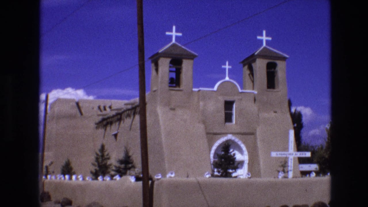 Adobe church front with bell towers, roof crosses, arched doorway, foreground wall, and utility line.