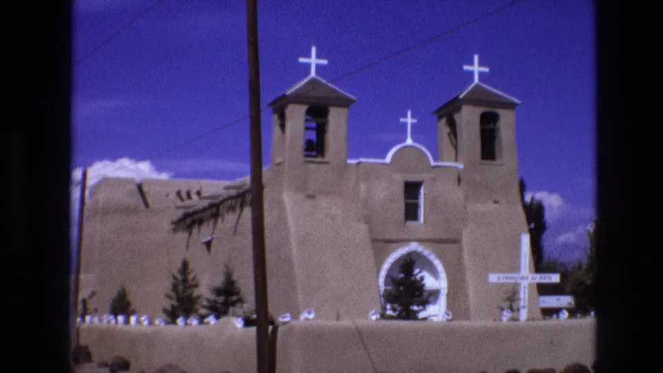 1973 New Mexico 8mm frame of the adobe church front, with two bell towers, several white crosses, a central arched entrance, a foreground wall, and a visible utility line across the sky.