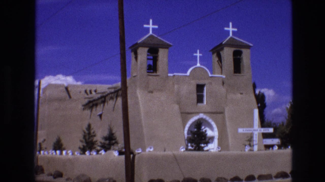 1973 New Mexico 8mm frame showing a wide exterior view of an adobe-style mission church with twin bell towers, white crosses, and a deep blue sky.