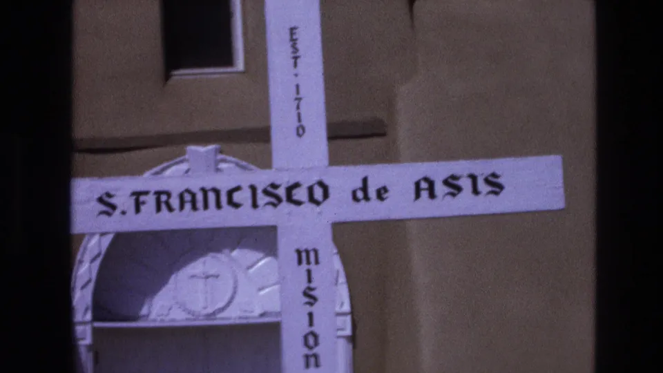 Close 1973 New Mexico 8mm view of the white S. FRANCISCO de ASIS mission cross marker against a tan adobe wall.