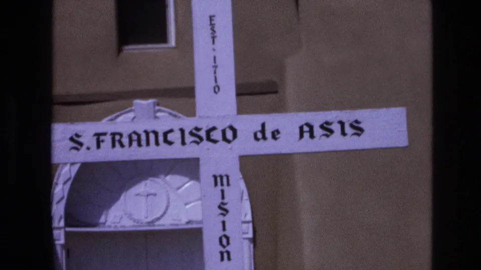 1973 New Mexico 8mm frame tightly composed on the S. FRANCISCO de ASIS cross marker, with EST. 1710 text and mission doorway details behind it.