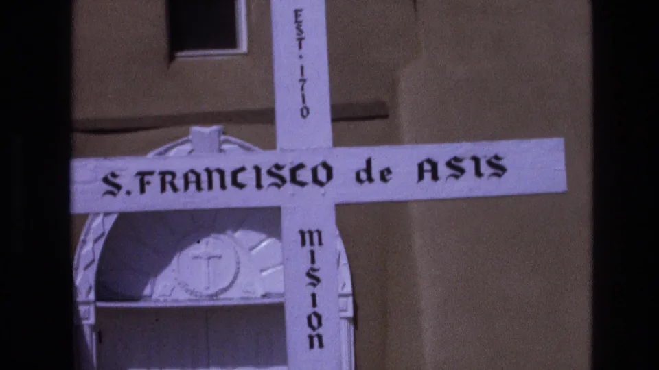 Final 1973 New Mexico 8mm frame holding on the S. FRANCISCO de ASIS cross sign, with a tan mission wall and arched entrance detail in the background.