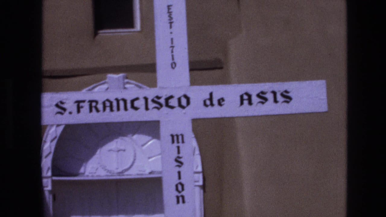 1973 New Mexico 8mm frame showing a close view of the white S. Francisco de Asis Mission cross with EST. 1710 and mission lettering against an adobe wall.