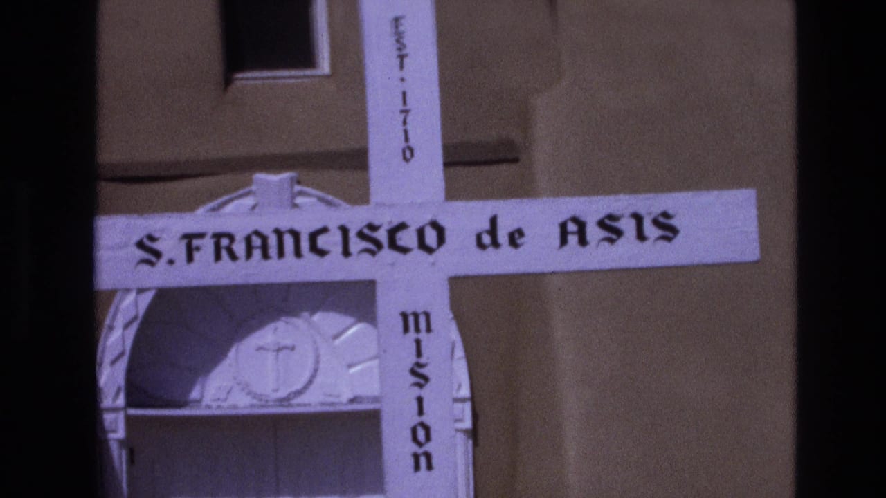1973 New Mexico home-movie frame with the mission cross centered near an adobe facade, including readable S. Francisco de Asis and EST. 1710 markings.