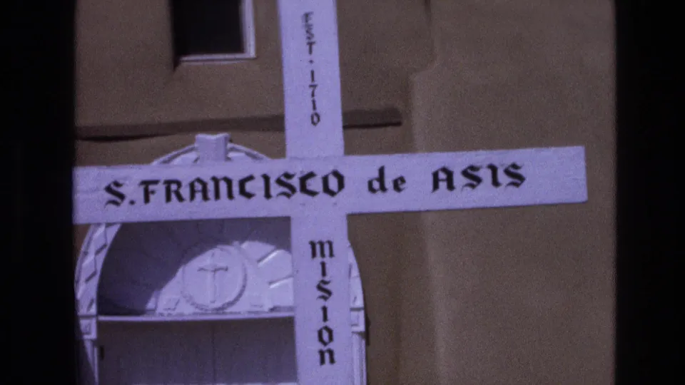 1973 New Mexico home-movie frame with the mission cross centered near an adobe facade, including readable S. Francisco de Asis and EST. 1710 markings.