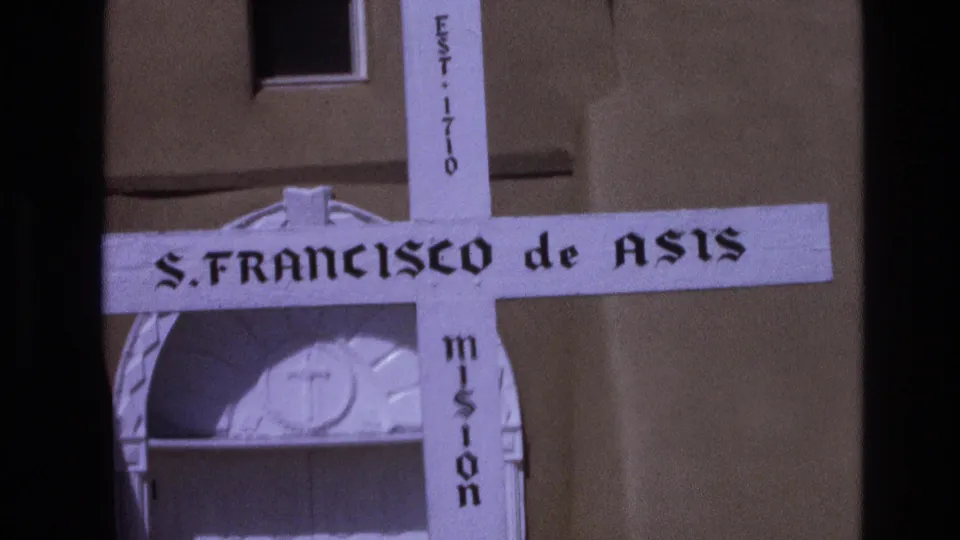 1973 New Mexico 8mm frame showing the white S. Francisco de Asis Mission cross below a small dark window, with mission text visible on the vertical beam.