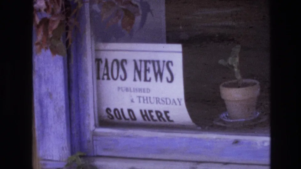 1973 New Mexico home-movie frame showing a storefront-style window display with a readable Taos News sign, Thursday publication note, and small potted plant.