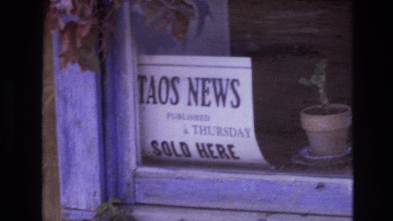 1973 New Mexico 8mm frame showing a white Taos News newspaper sign in a weathered wooden window beside a small potted plant.