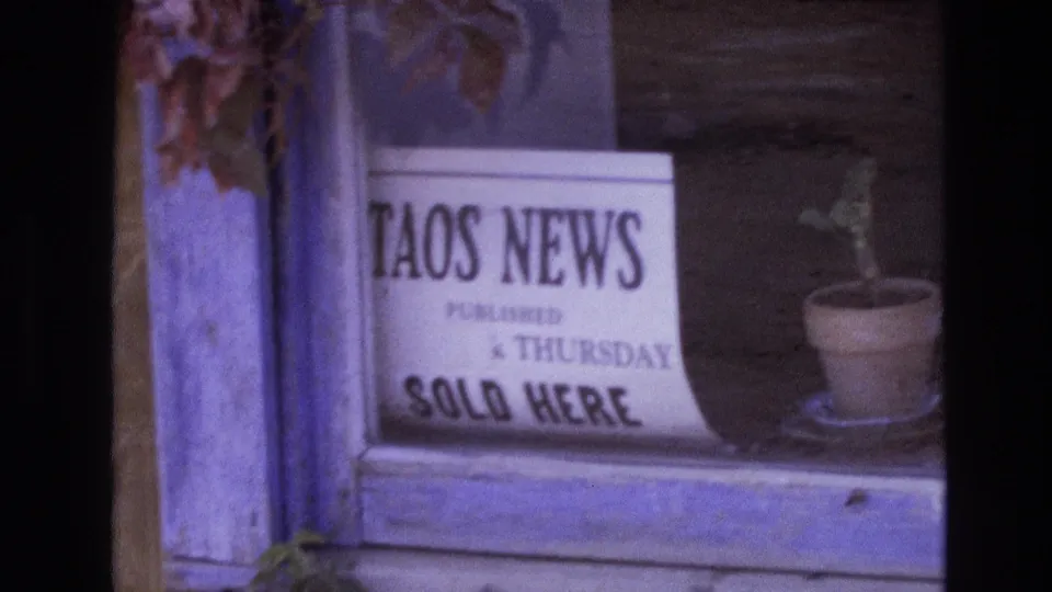 1973 New Mexico 8mm frame showing a white Taos News newspaper sign in a weathered wooden window beside a small potted plant.