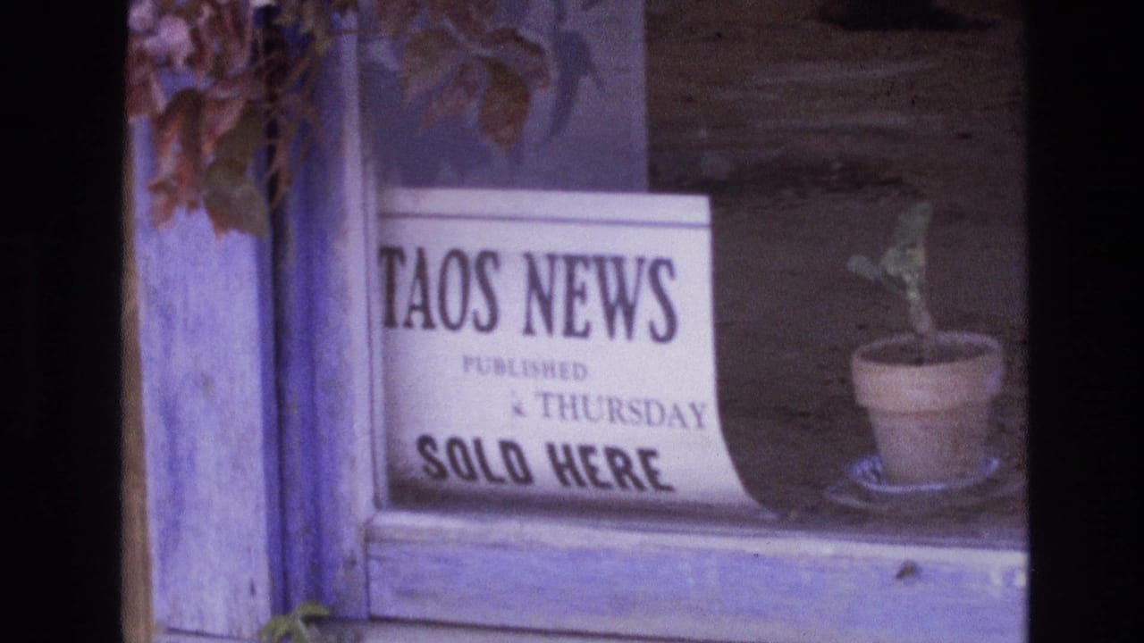 White Taos News sign and potted plant seen through a wooden window frame.