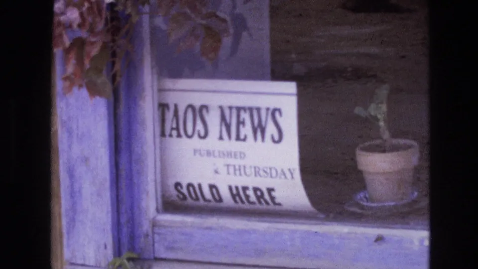 1973 New Mexico home-movie frame with the Taos News sign centered in a window and a small plant pot on the sill to the right.