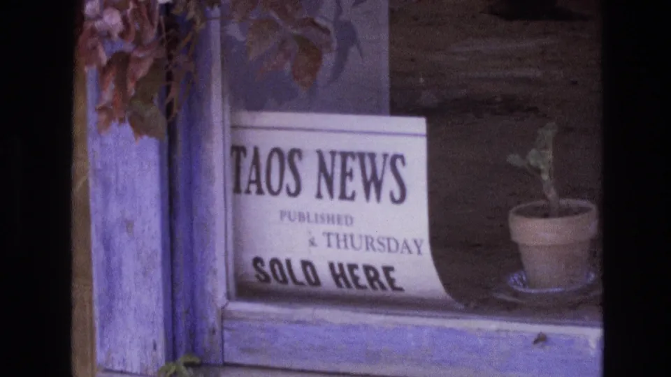 Soft 8mm frame from New Mexico showing the Taos News sold-here sign cropped at the left edge, with a small potted plant visible on the right.