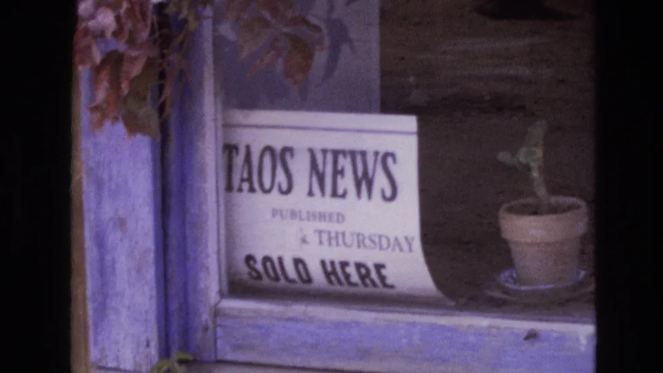 Final 1973 New Mexico 8mm frame holding on the Taos News window sign, potted plant, rough wood frame, and muted purple film color.