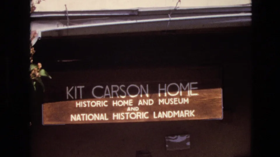 1973 New Mexico 8mm frame showing the Kit Carson Home historic home and museum National Historic Landmark sign mounted on a dark wooden exterior.