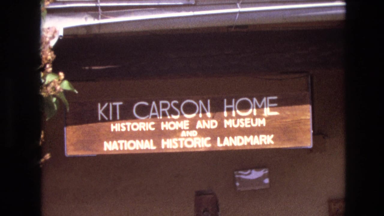 1973 New Mexico home-movie frame showing a close view of the wooden Kit Carson Home Historic Home and Museum National Historic Landmark sign mounted on an adobe-style wall.