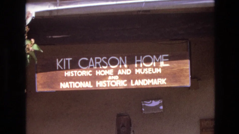 1973 New Mexico home-movie still showing the Kit Carson Home sign in a slightly soft close frame, with the adobe-style wall, roof edge, and dark film border around it.