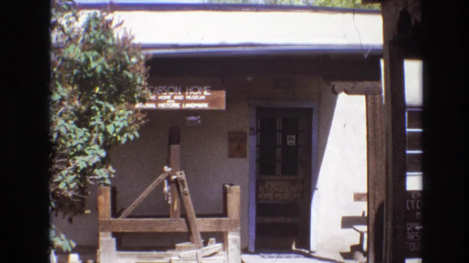 1973 New Mexico frame pulling back to show the Kit Carson Home sign above an adobe-style entrance area, with greenery, a dark doorway, and wooden gate or fence elements in front.