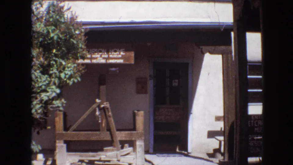 1973 New Mexico visitor footage showing the front of the Carson Home and Museum with a wooden sign, shaded doorway, tree, and rustic well structure.