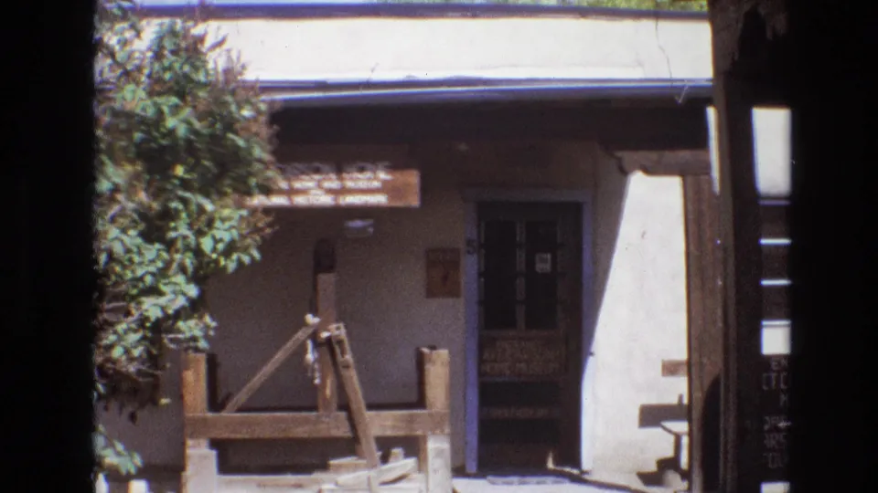 1973 New Mexico home-movie frame showing the Carson Home and Museum facade with a rustic wooden well structure, shaded entrance, and leafy tree at left.