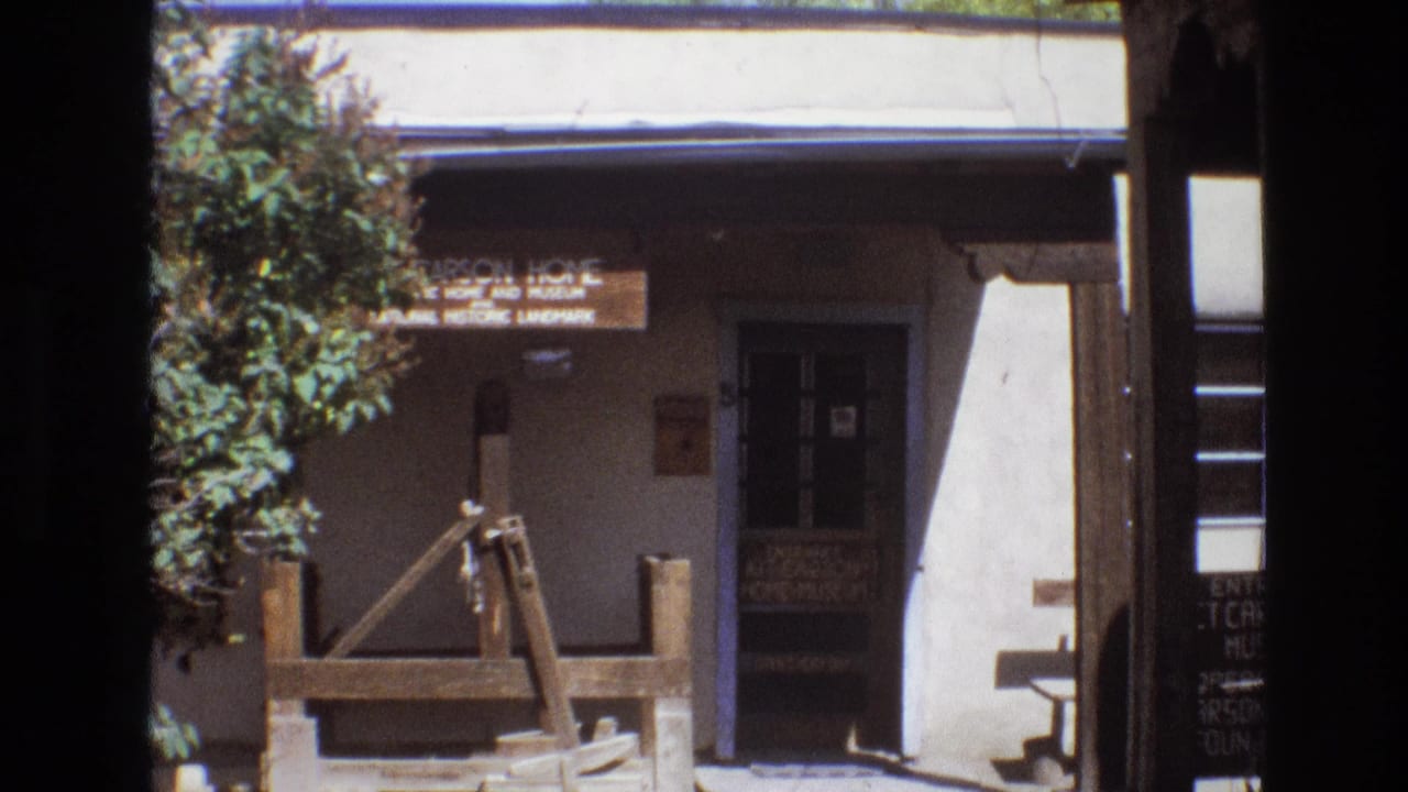 1973 New Mexico 8mm frame showing the exterior of a historic adobe museum with a shaded entrance, sign, trees, and rustic wooden display pieces.