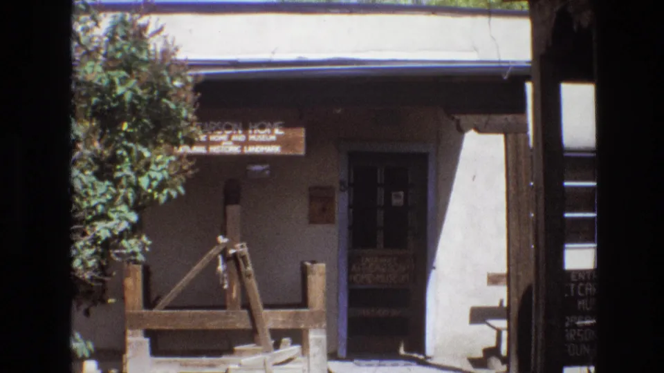 1973 New Mexico 8mm frame showing the exterior of a historic adobe museum with a shaded entrance, sign, trees, and rustic wooden display pieces.