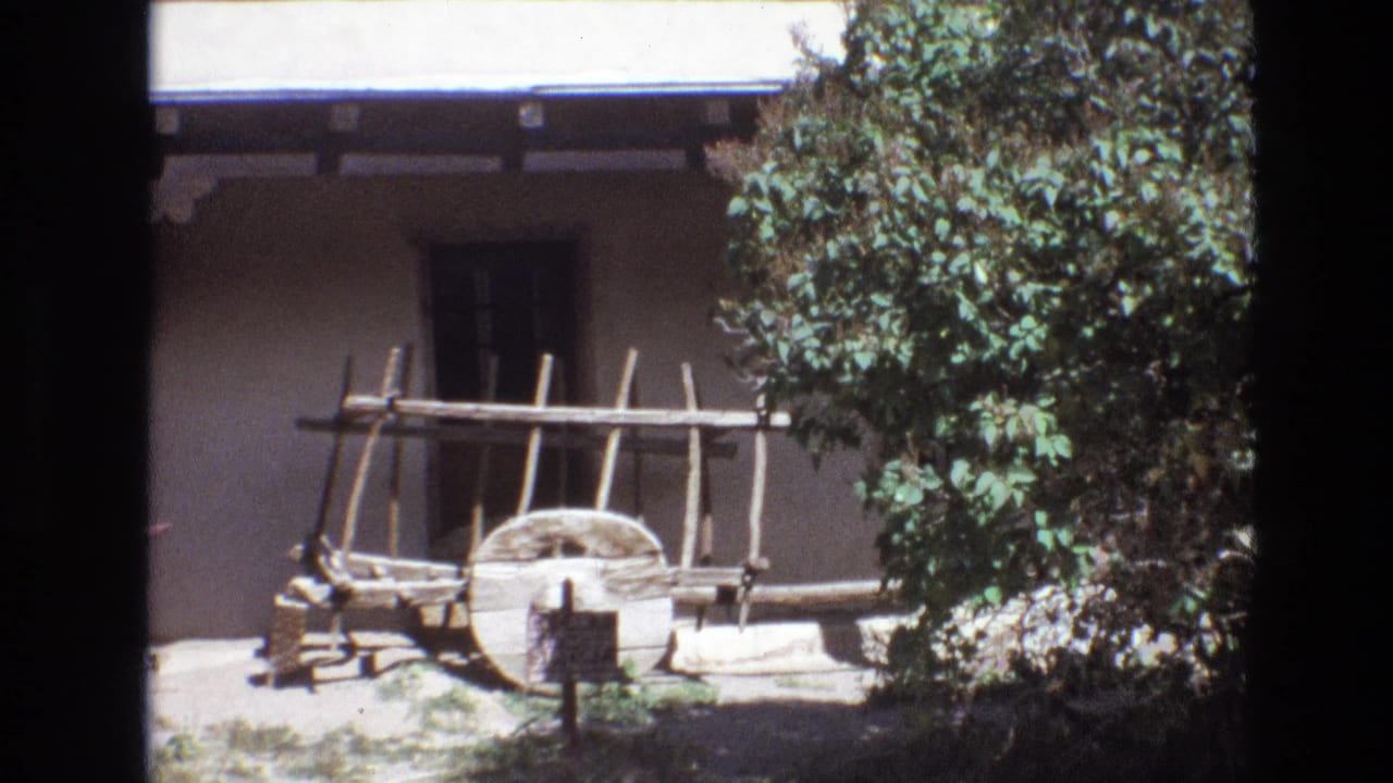 Wooden cart and rack displayed outside an adobe building, partly framed by tree foliage.