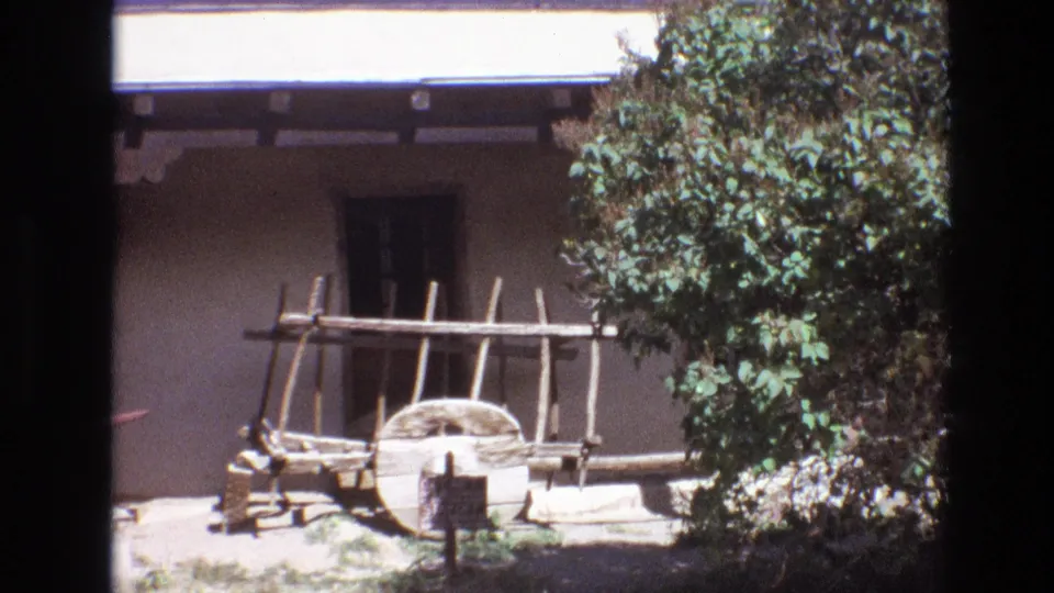 1973 New Mexico 8mm frame showing the rustic wooden cart display against an adobe museum exterior, with trees and shrubs along the edge of the frame.