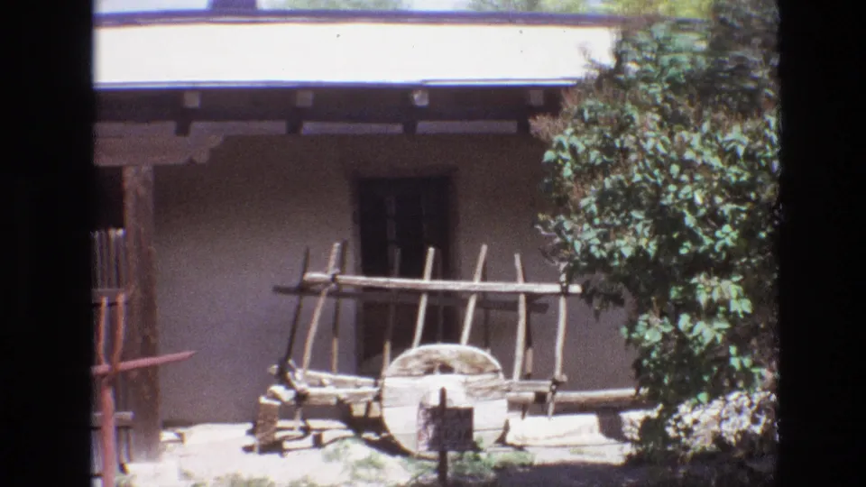 1973 New Mexico 8mm frame of the museum exterior with a wooden cart display, small window, adobe wall, and leafy tree in strong sun.