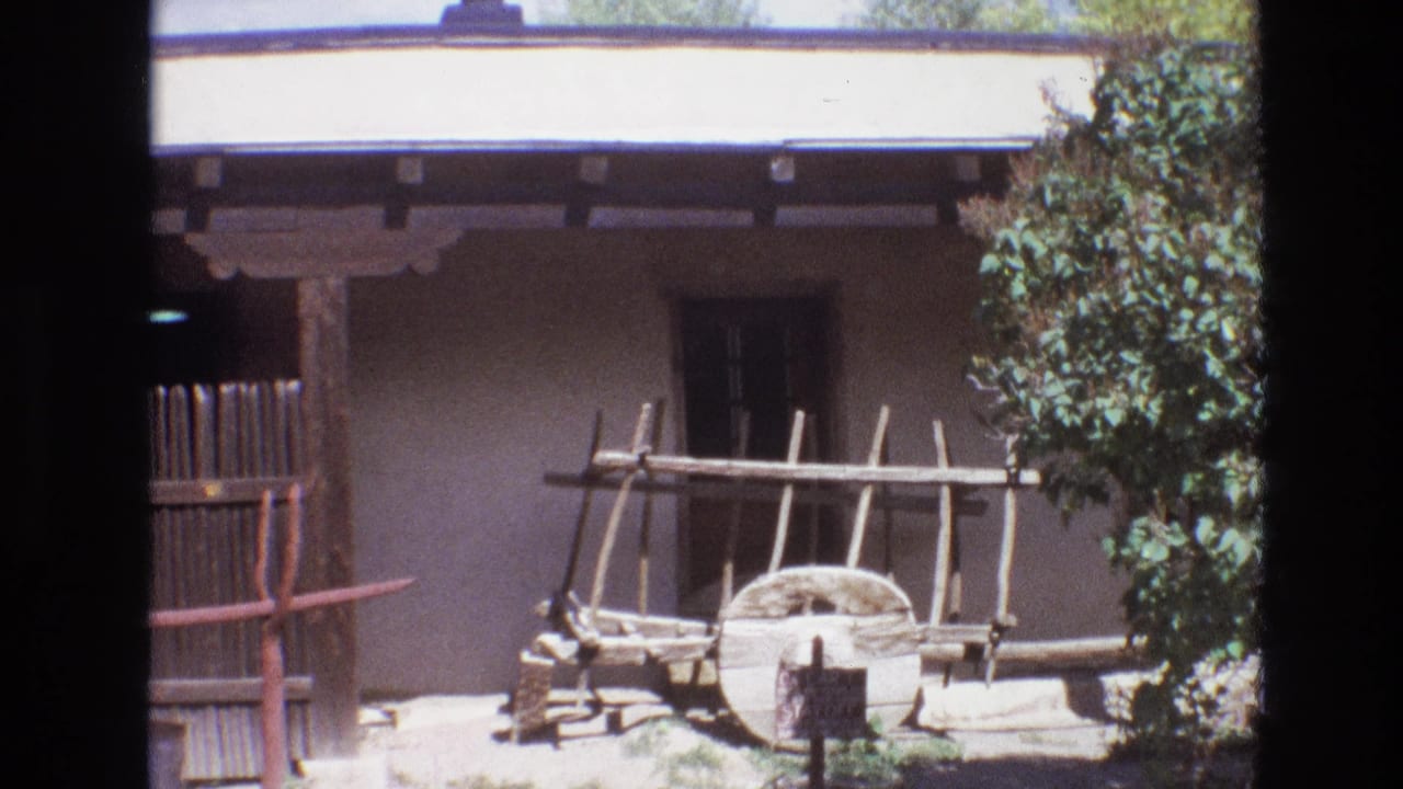 1973 New Mexico 8mm frame showing an adobe exterior with shaded porch beams, a vertical wooden fence, a wagon wheel, a wooden rack, and partial greenery at the right edge.