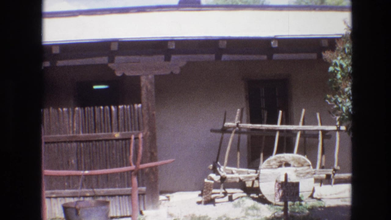 1973 New Mexico frame with an adobe wall, shaded porch, vertical wooden fence, wagon wheel, wooden rack, red-brown rail, and small sign in bright daylight.