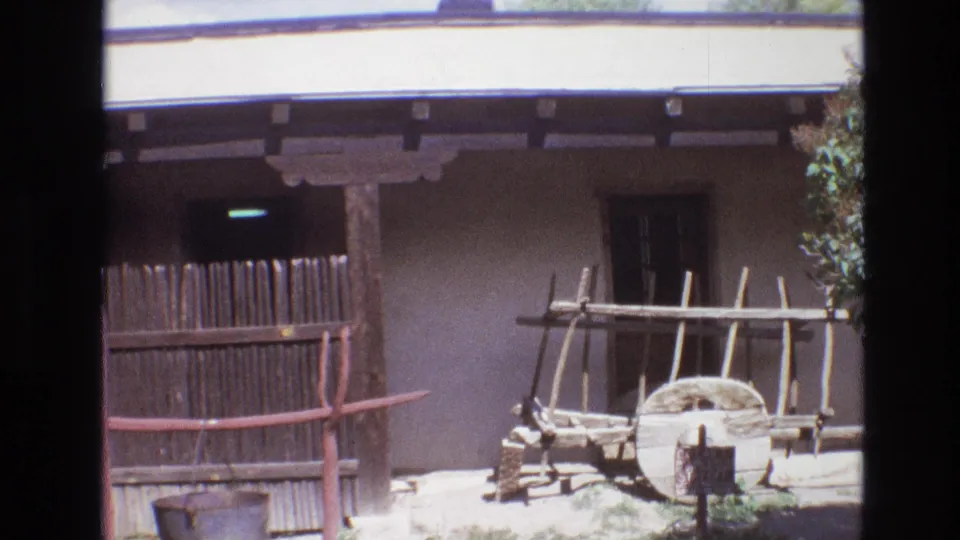 1973 New Mexico frame with an adobe wall, shaded porch, vertical wooden fence, wagon wheel, wooden rack, red-brown rail, and small sign in bright daylight.