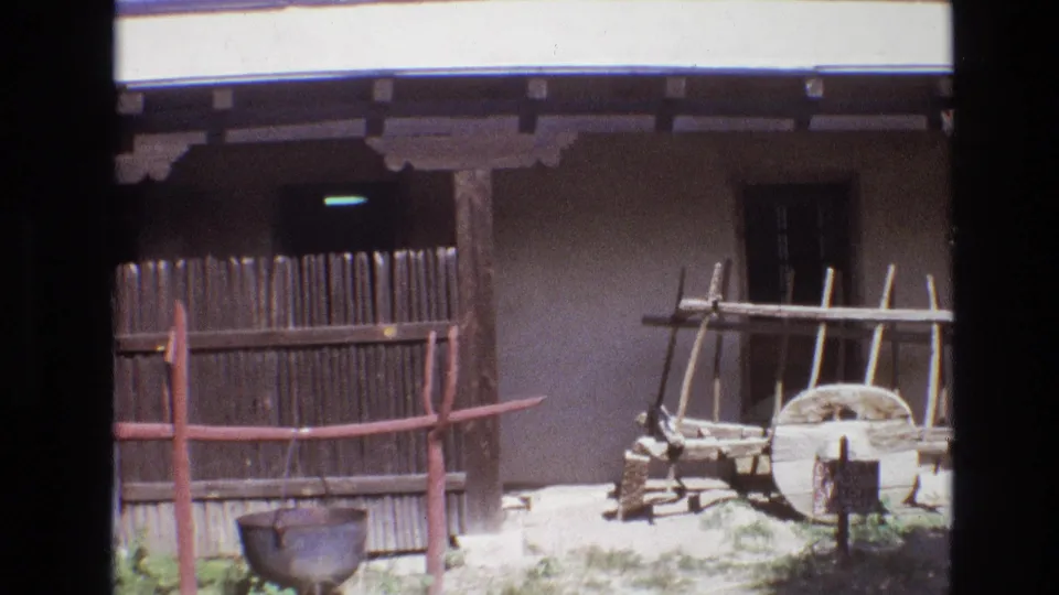 1973 New Mexico 8mm frame showing a closer courtyard view with a vertical wooden fence, red-brown rail, hanging pot, adobe wall, wagon wheel, and wooden rack.