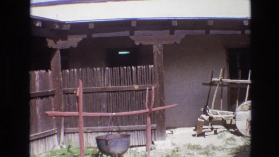 1973 New Mexico frame focused on the wooden fence and hanging pot, with the adobe wall, shaded porch, partial wagon wheel, and wooden rack visible to the right.