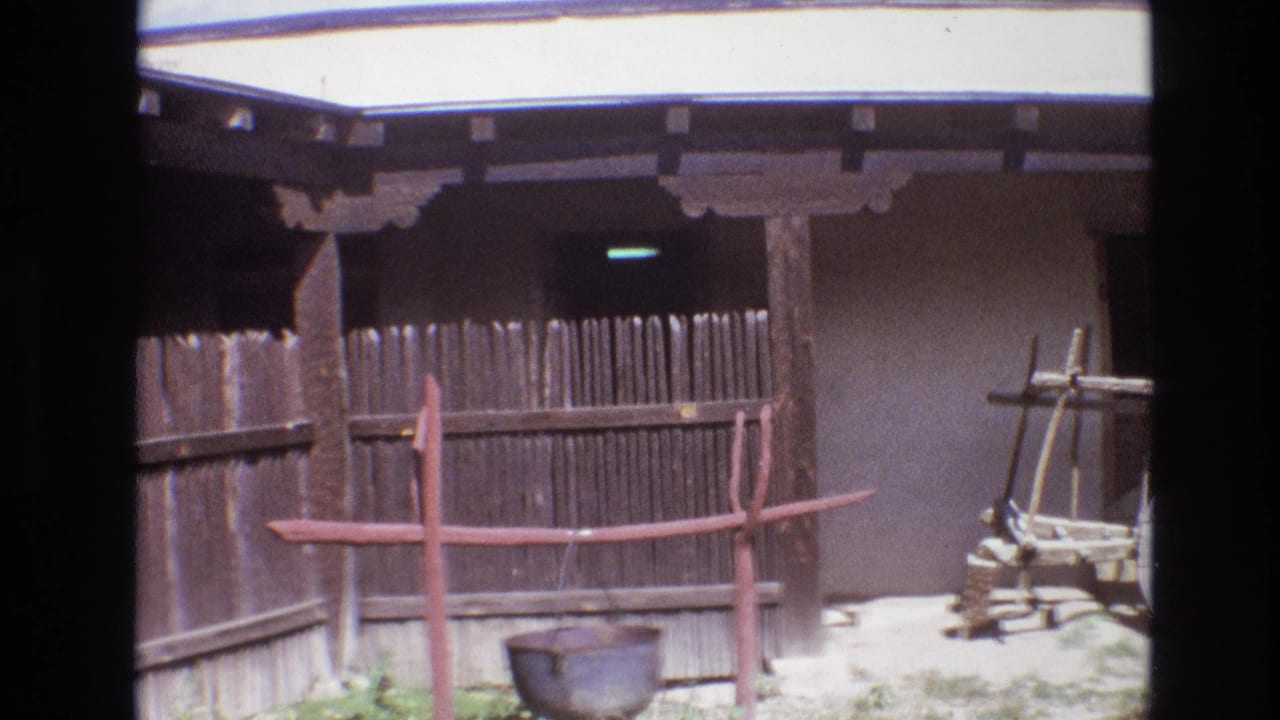 1973 New Mexico 8mm frame showing a rustic wooden fence, red-painted support frame, hanging metal pot, and adobe-style exterior wall in a sunlit outdoor courtyard.