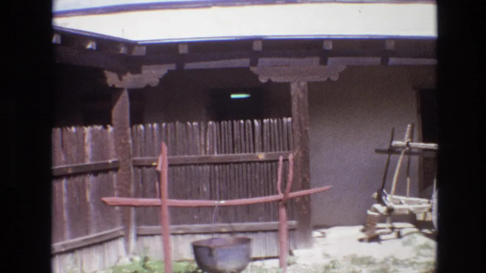 1973 New Mexico 8mm frame showing a rustic wooden fence, red-painted support frame, hanging metal pot, and adobe-style exterior wall in a sunlit outdoor courtyard.