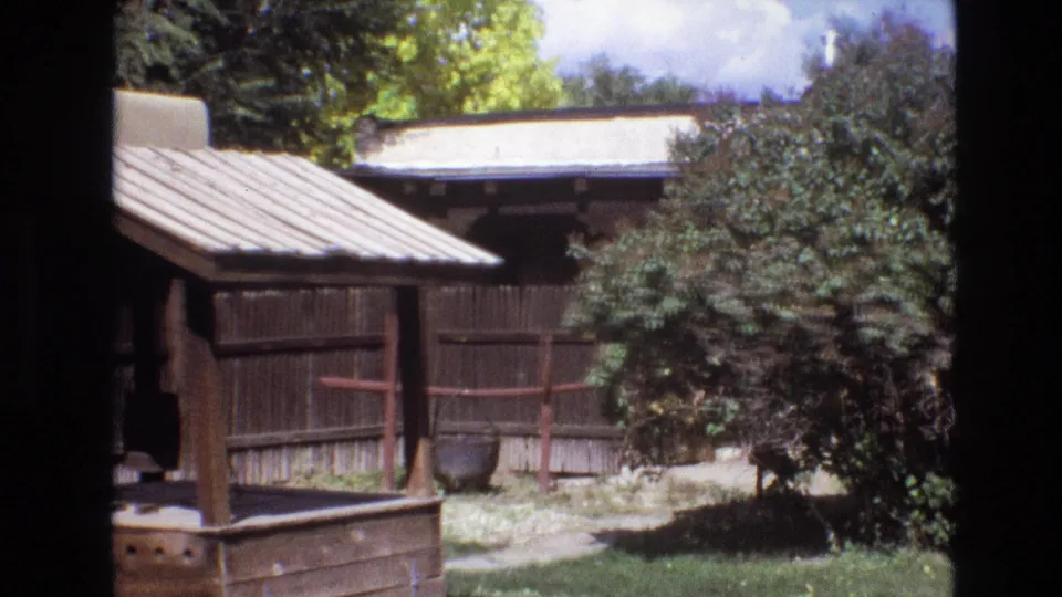1973 New Mexico 8mm frame with a small covered wooden well-like structure, plank fencing, shrubs, grass, and a dark round pot near the fence.