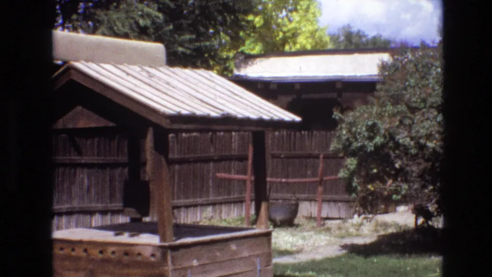 1973 New Mexico 8mm frame showing the sloped roof and posts of a small wooden structure beside a weathered plank fence, shrubs, and patchy grass.