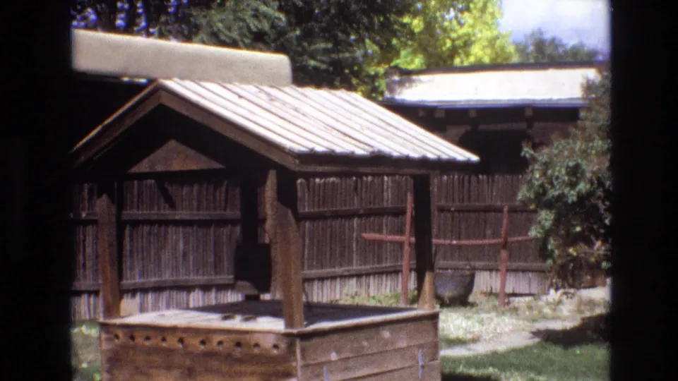 1973 New Mexico 8mm frame showing a rustic covered wooden structure in the foreground, vertical plank fencing, trees, shrubs, grass, and a dark pot near the fence.