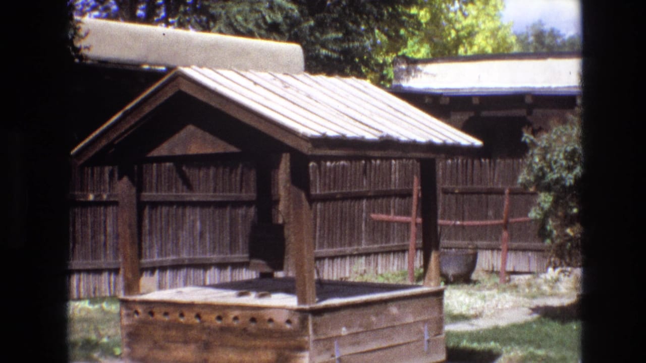 A wooden well-like structure stands in a fenced yard, with trees and low buildings visible in 1973 New Mexico home-movie footage.