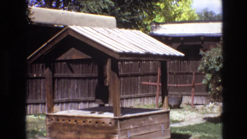 A wooden well-like structure stands in a fenced yard, with trees and low buildings visible in 1973 New Mexico home-movie footage.