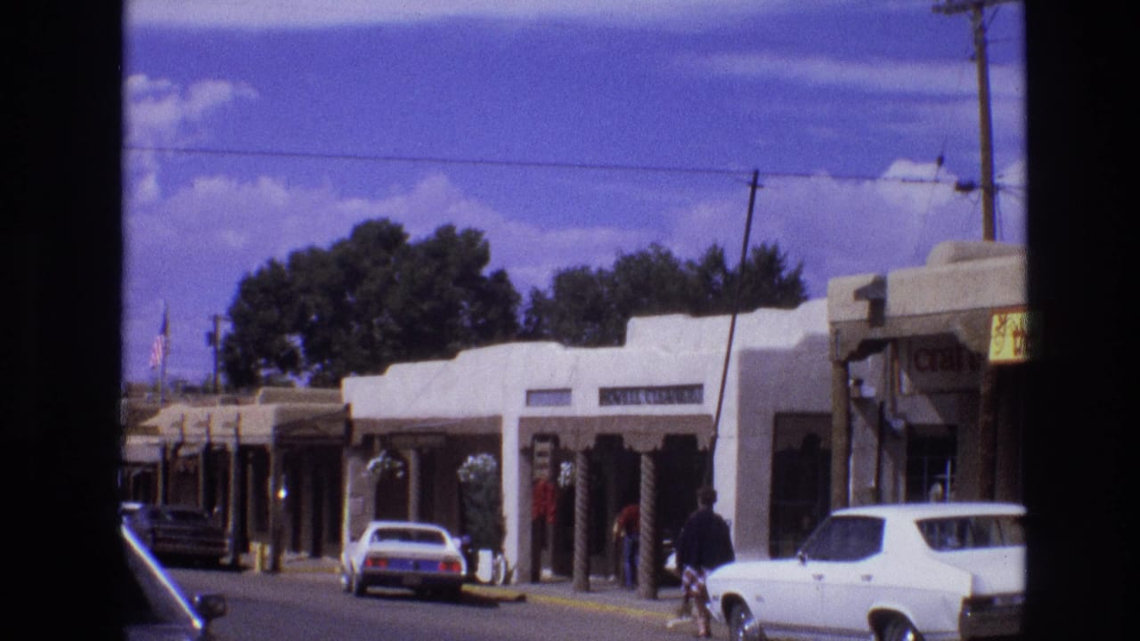 Adobe-style storefronts with parked cars, pedestrians, utility lines, and a distant flag.