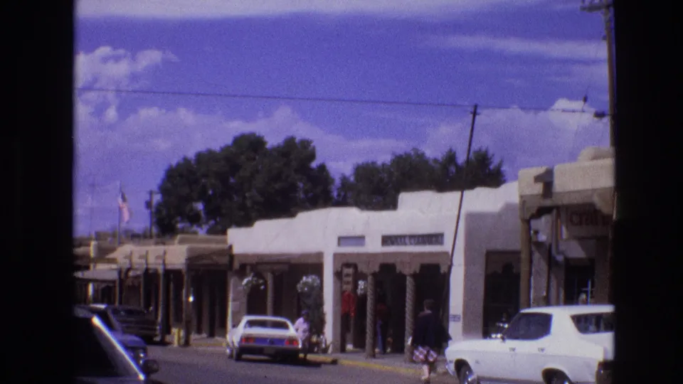 A 1973 New Mexico main street view shows adobe-style shops, a white parked car, pedestrians near storefronts, utility poles, and a bright clouded sky.