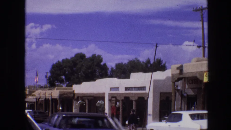 The street scene continues with adobe-style shopfronts, a white vehicle at the curb, pedestrians on the sidewalk, a dark car in the foreground, utility lines, and trees under a blue sky.