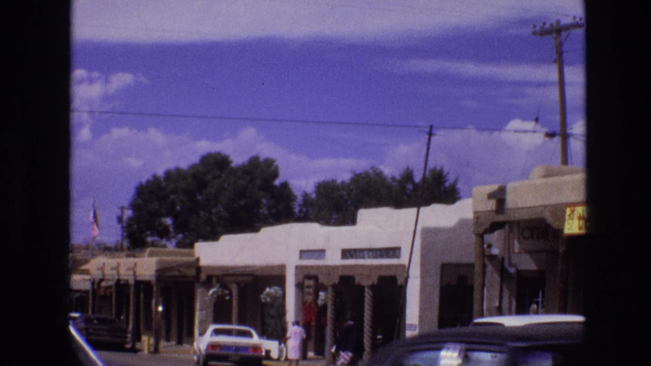 1973 New Mexico home-movie frame showing a sunlit row of Southwestern adobe-style storefronts, parked cars, a few pedestrians, utility wires, and dark 8mm frame borders.