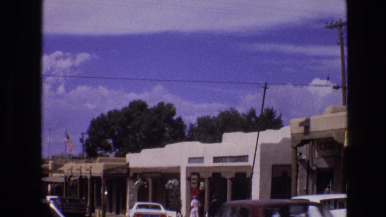 Small-town shopping strip with adobe-style buildings, parked cars, pedestrians, and overhead wires.