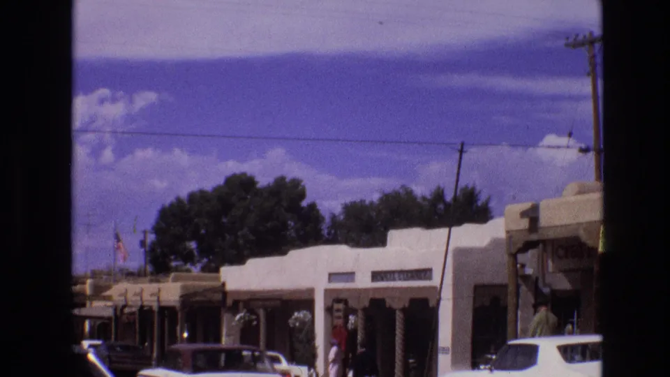 1973 New Mexico home-movie frame showing a commercial row of Southwestern storefronts, shaded awnings, parked cars, pedestrians, utility poles, and saturated 8mm color.