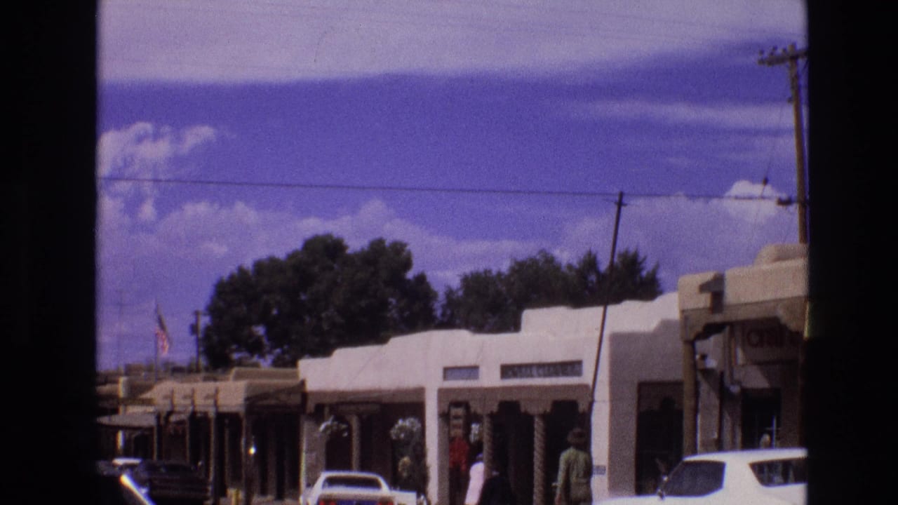 1973 New Mexico 8mm frame showing low adobe-style storefronts, several older cars, a few pedestrians, utility lines, and mountains under a bright sky.