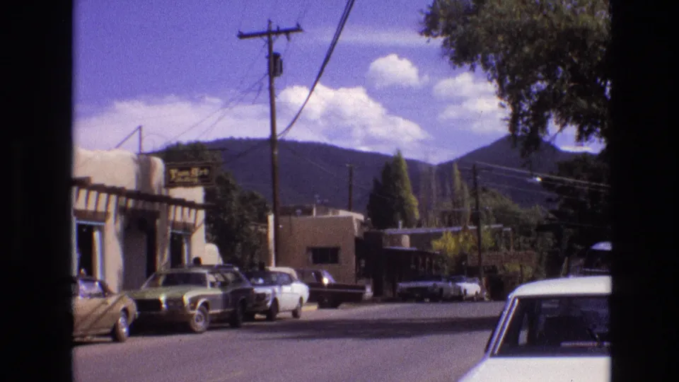 1973 New Mexico street frame with older parked cars, a business sign, utility lines crossing the scene, trees, white clouds, and a mountain range beyond town buildings.