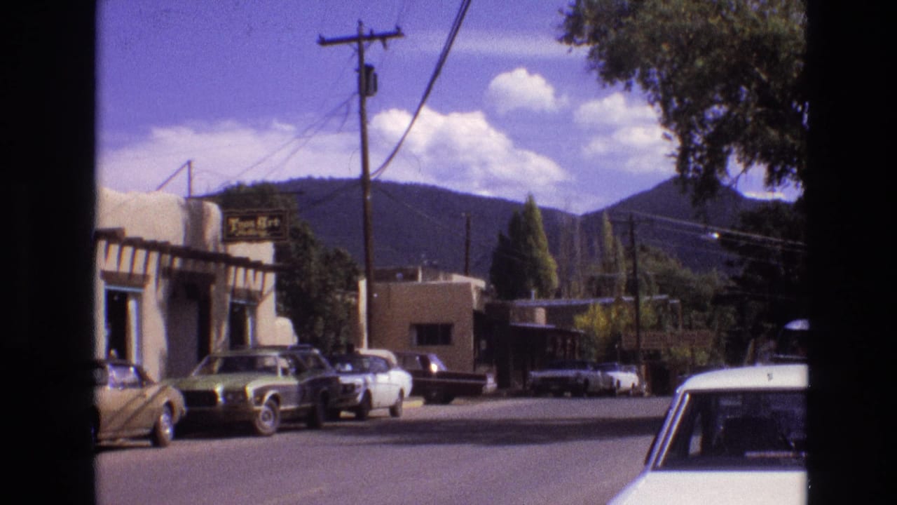 Parked cars and low buildings along a sunny street with utility wires and mountains behind them.