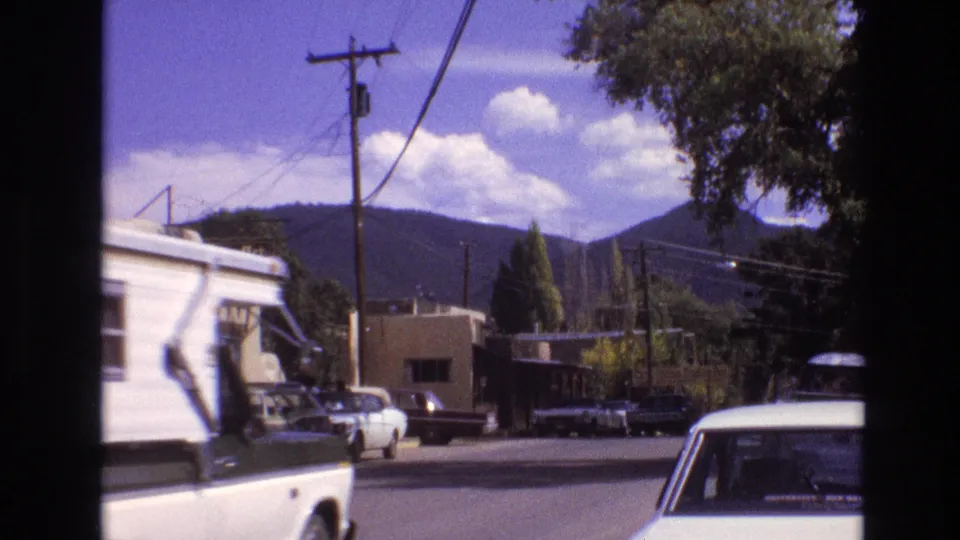 1973 New Mexico home-movie frame with a white camper entering the street view, parked cars along the curb, utility lines overhead, and mountains in the background.